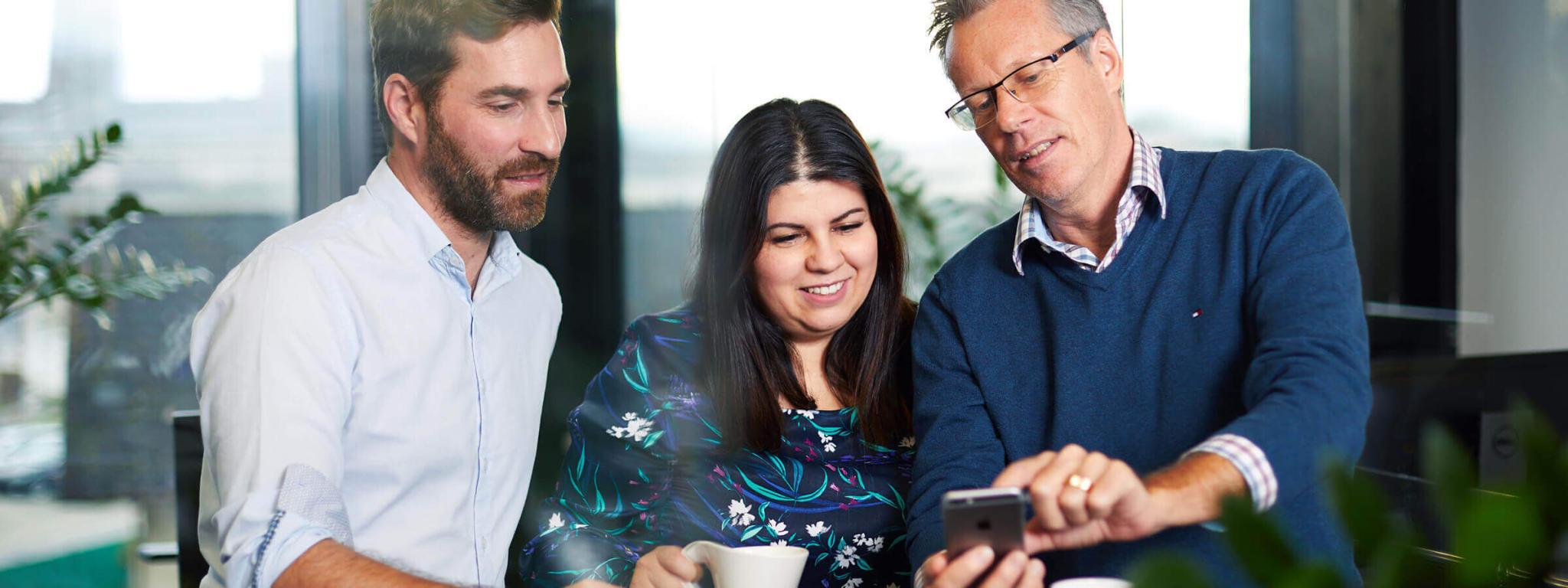 Zwei Männer und eine Frau sehen auf ein Handy und trinken Kaffee